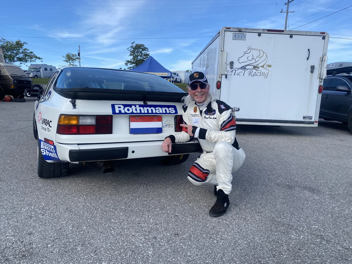 A driver in a white racing suit beside the number 9 Porsche and team trailer